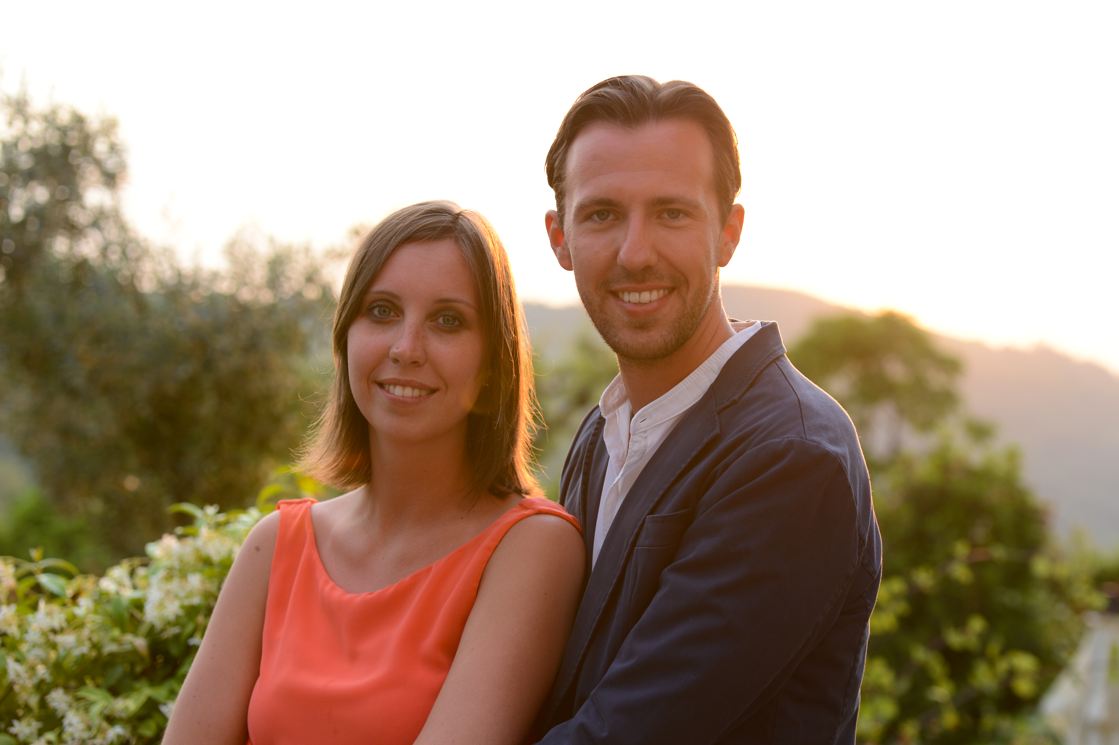A smiling couple poses together outdoors, with the man embracing the woman from behind.