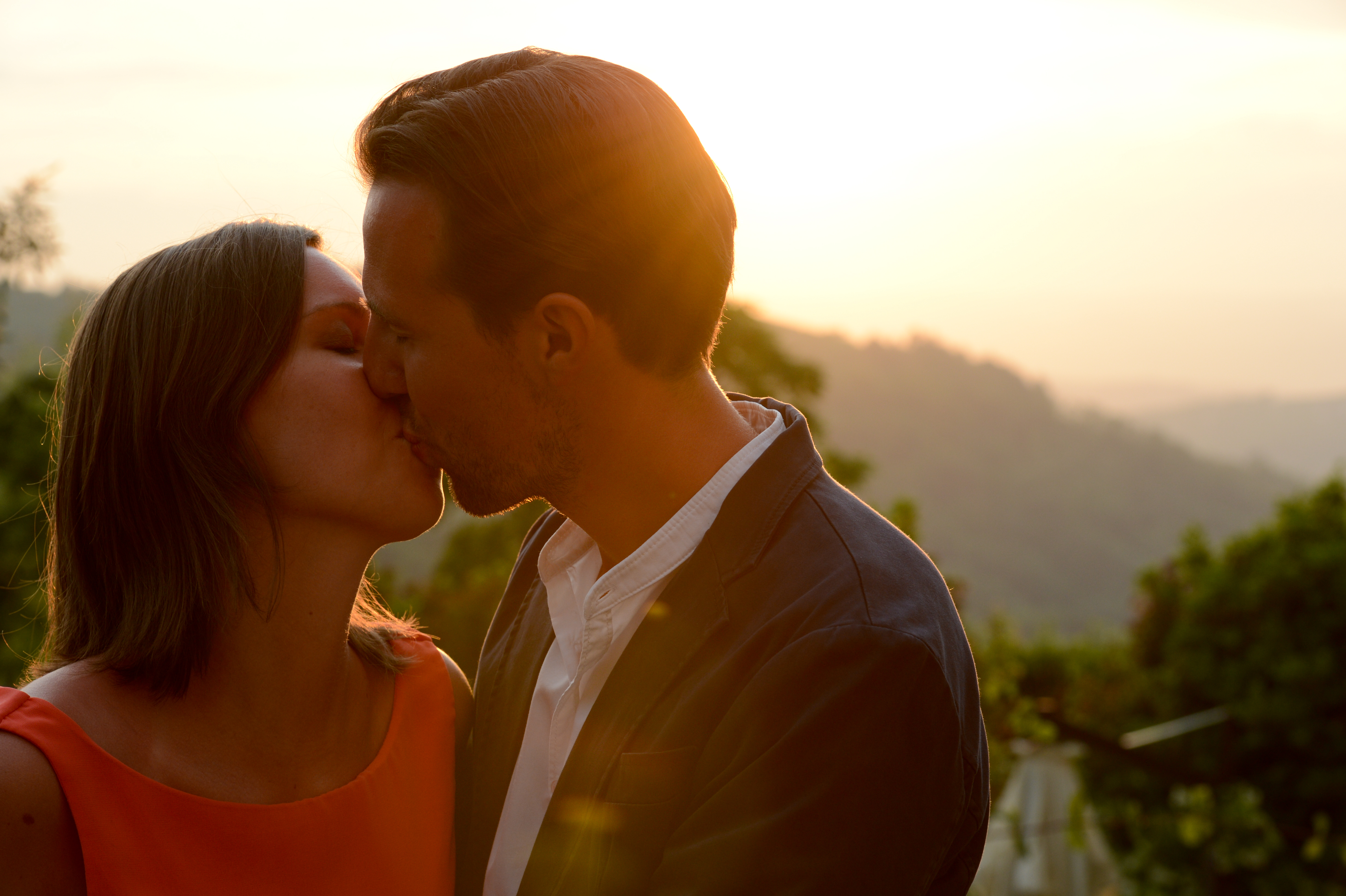 A couple shares a kiss outdoors with a sunset in the background.
