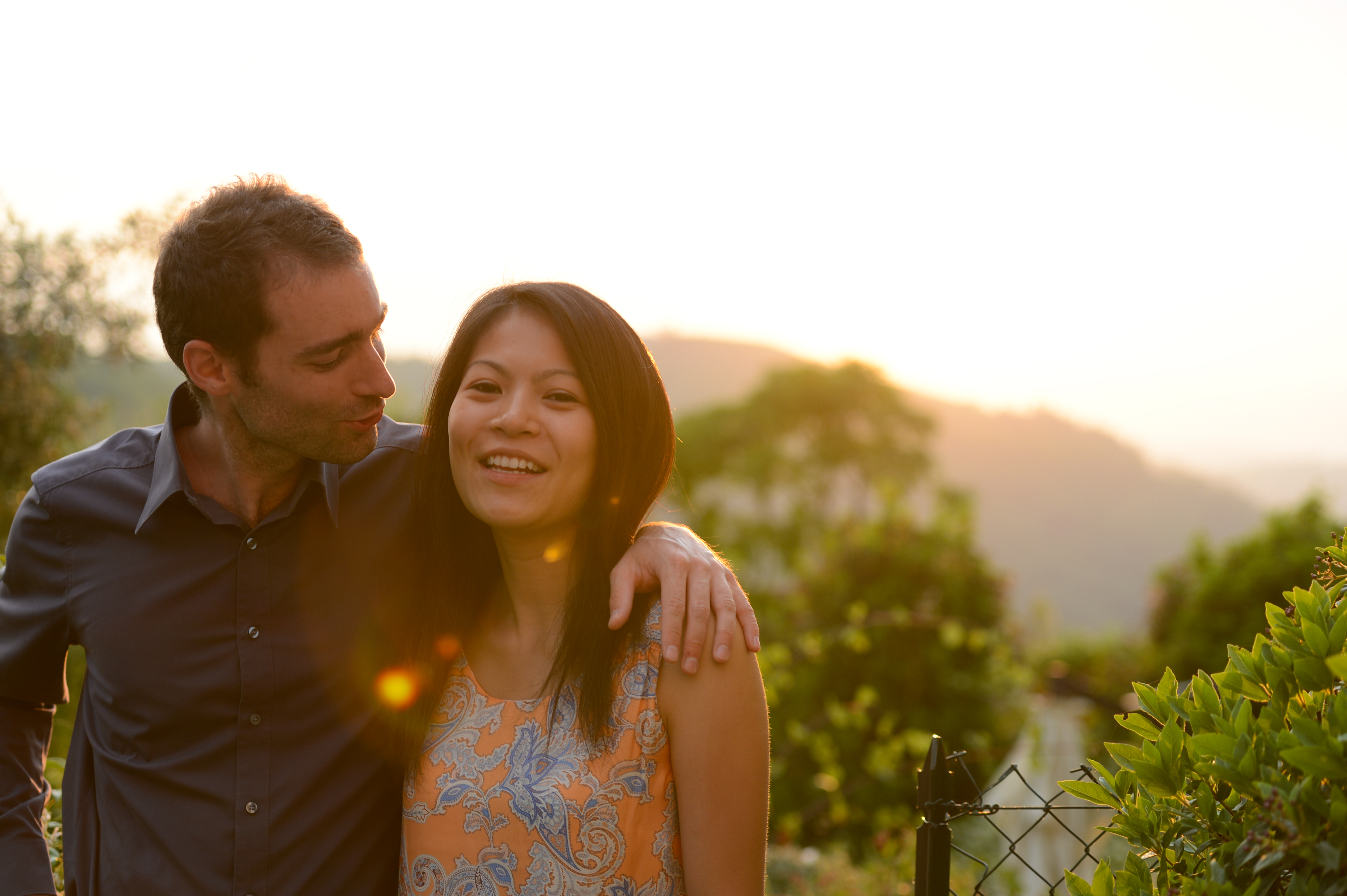 A smiling woman and a man with his arm around her stand outdoors during sunset.