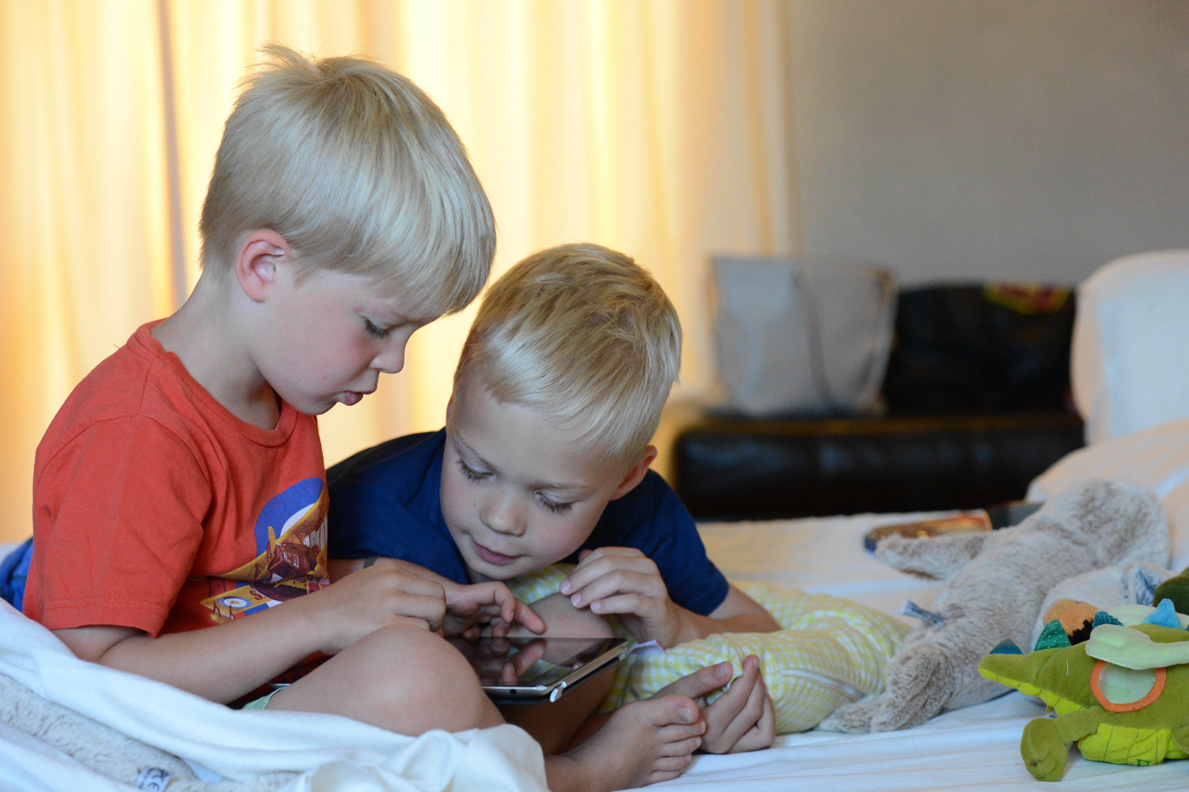 Two young boys sit on a bed, closely looking at a tablet together, surrounded by stuffed toys.