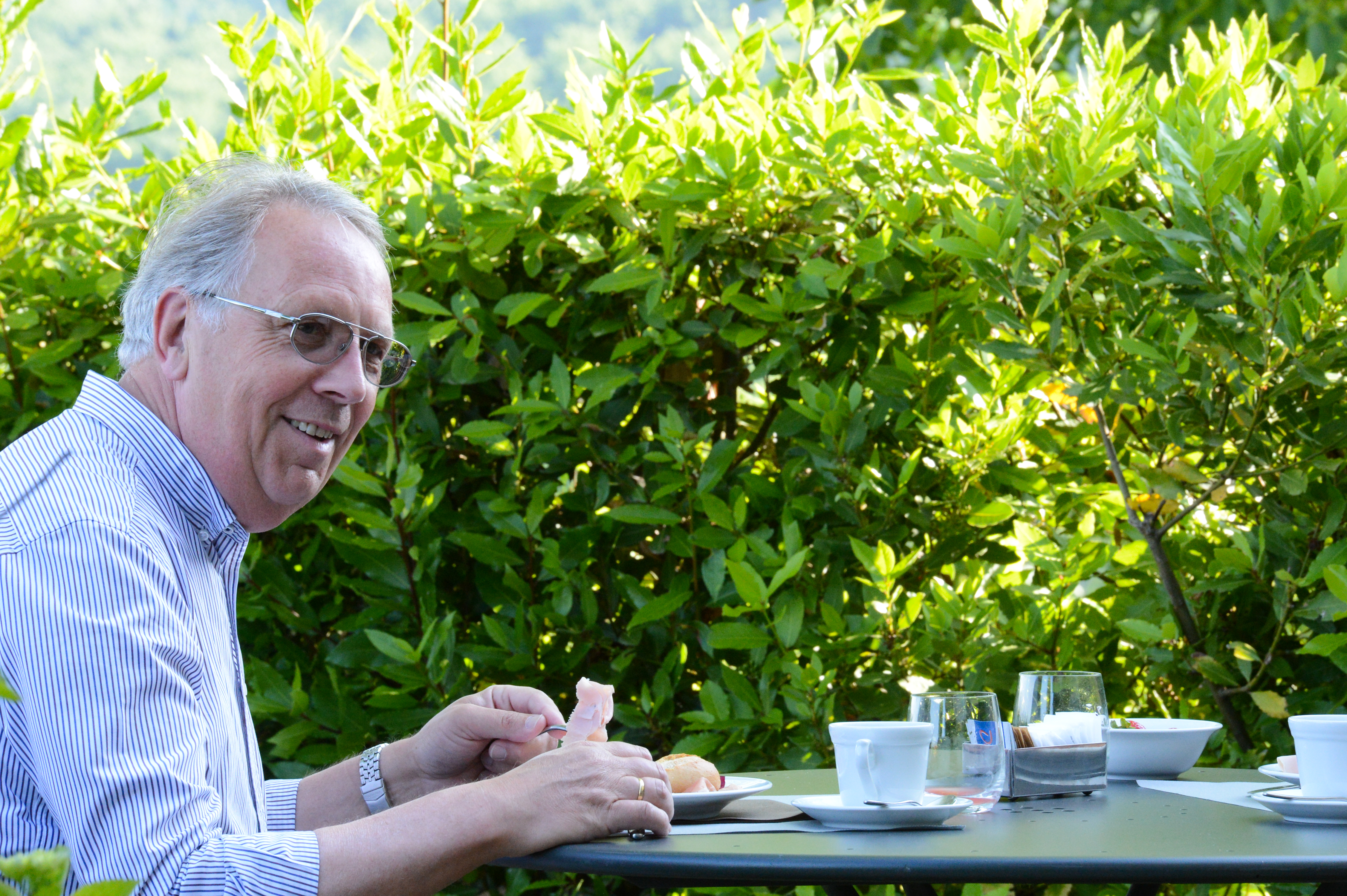 A man wearing glasses and a striped shirt smiles while eating at an outdoor table with food and drinks.