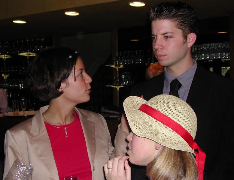 Three people in formal attire are engaged in conversation at a wedding reception, with glasses and decorations in the background.