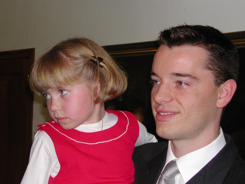 A man in a suit holds a young girl in a red dress as they both look to the side.