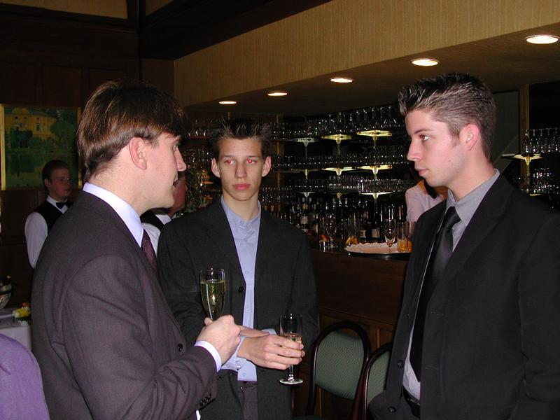 Three men in suits stand near a bar, holding champagne glasses and engaged in conversation at a wedding reception.