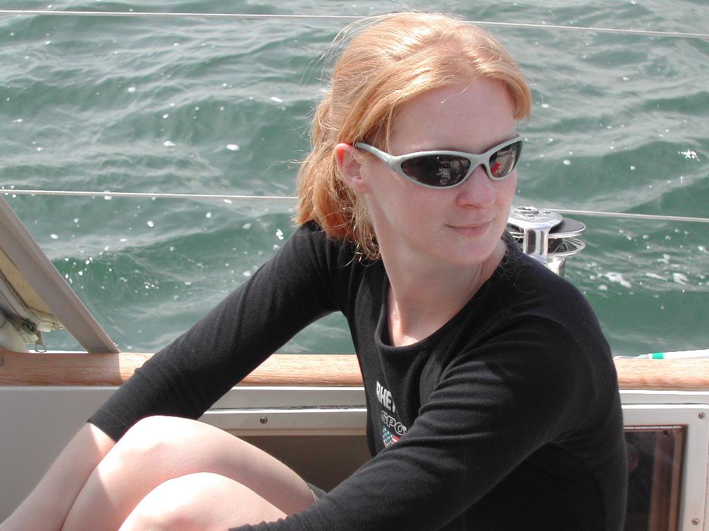 A woman wearing sunglasses and a black shirt sits on a boat, looking to the side with a relaxed expression.