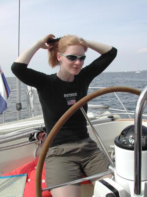 A woman wearing sunglasses sits at the helm of a sailboat, tying her hair back with both hands.