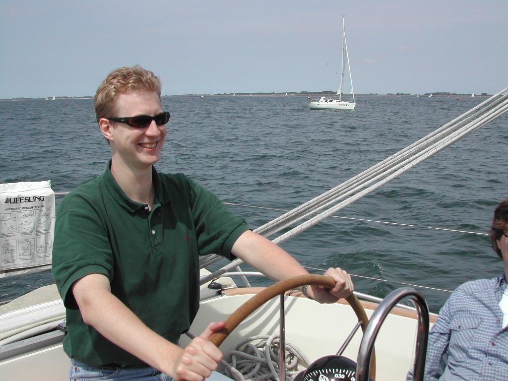 A man wearing sunglasses and a green shirt steers a sailboat, smiling.