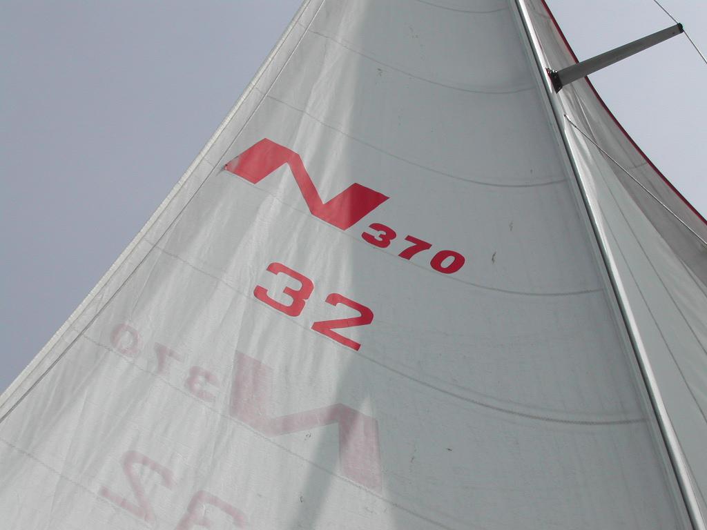 A close-up view of a sail with red markings, attached to a mast, against a cloudy sky.