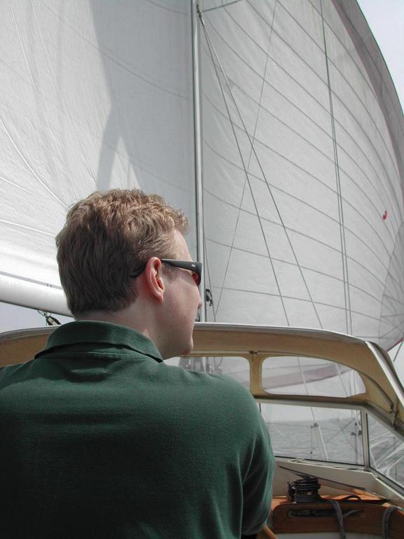 A man wearing sunglasses and a green shirt looks ahead while sailing on a boat with large white sails.