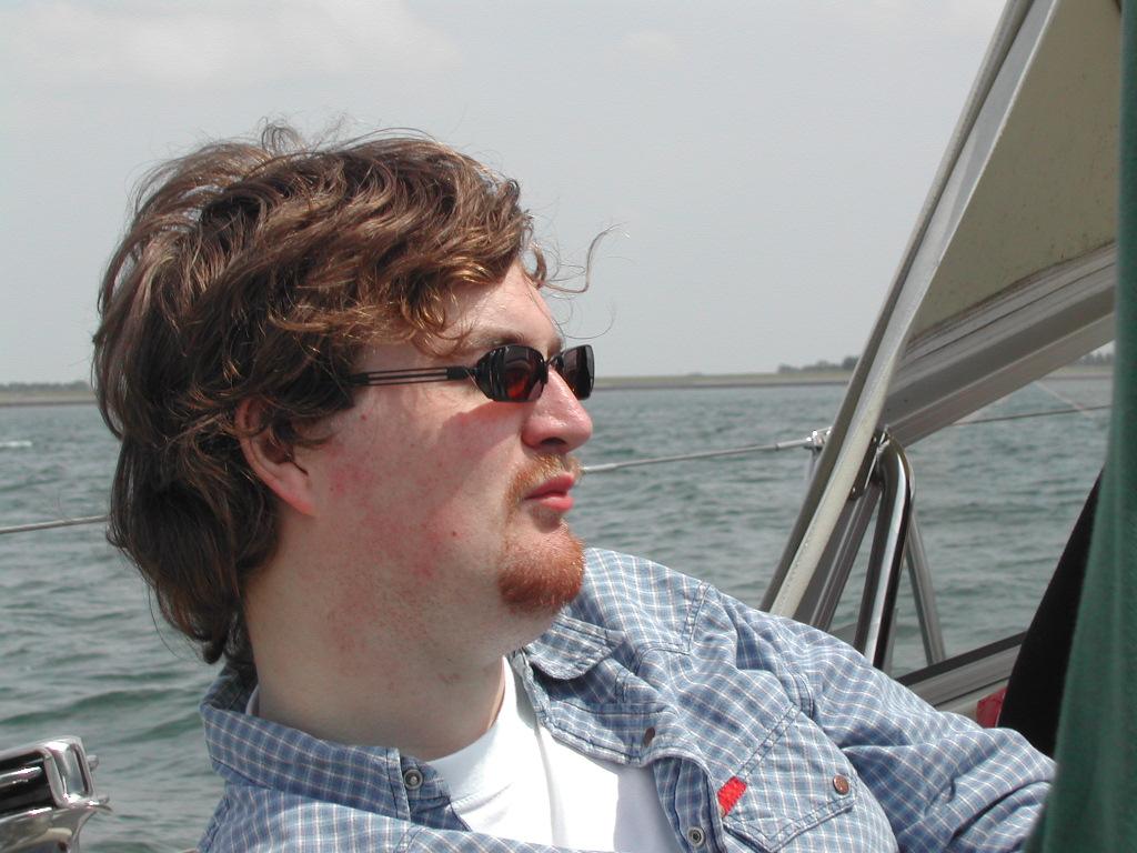 A man wearing sunglasses and a checkered shirt sits on a boat, looking out over the water.