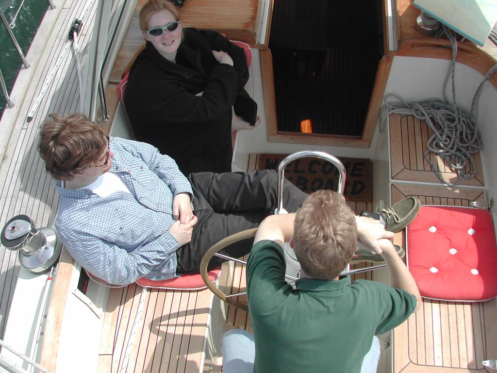 Three people are sitting on a boat, one steering while the others relax and talk.