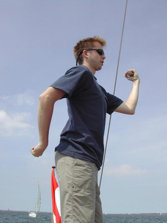 A person wearing sunglasses and a navy shirt flexes their arm while standing on a boat with a sail in view.