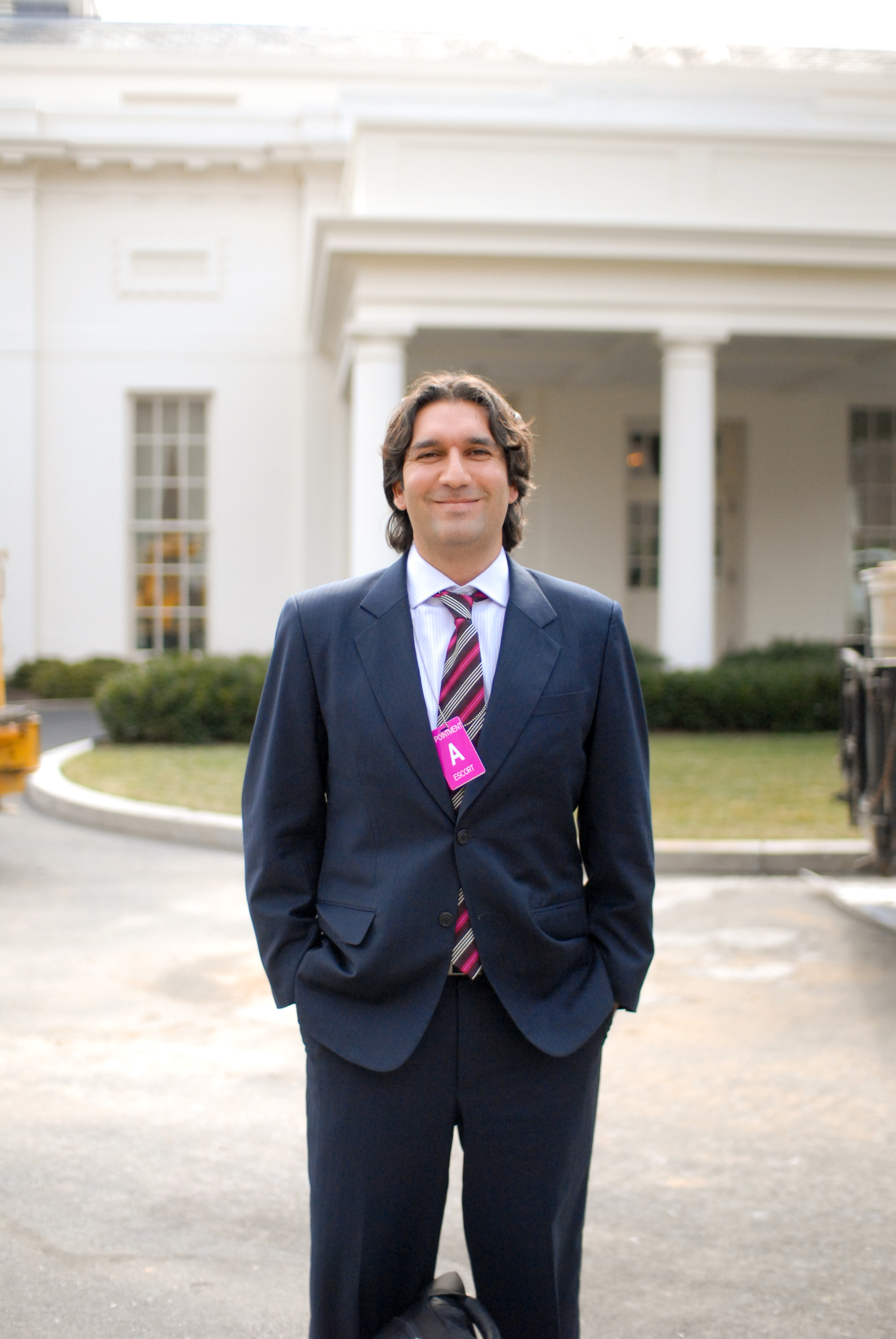 A man in a suit with a visitor badge stands outside the White House, smiling at the camera.
