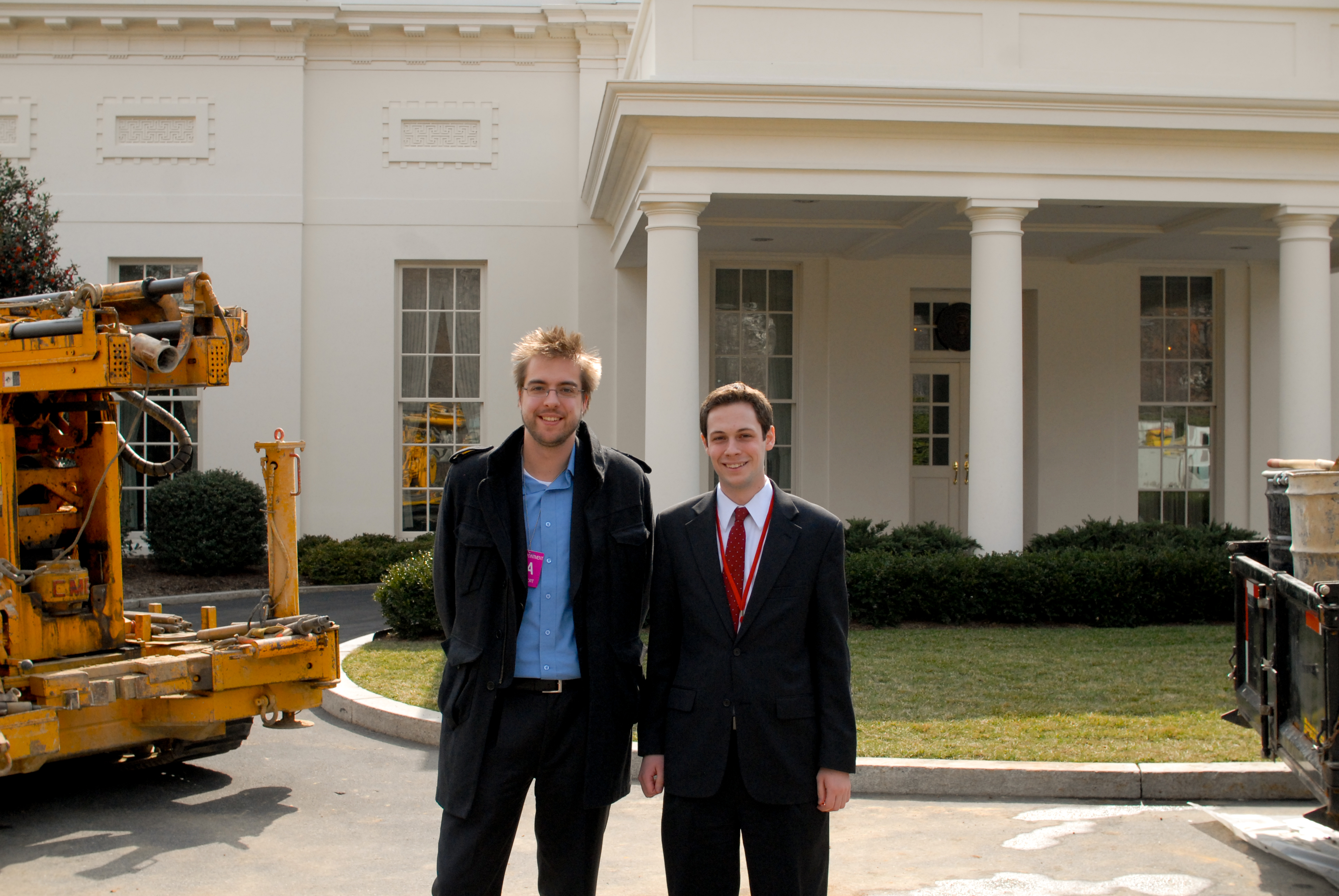 Two men stand smiling in front of the White House, wearing formal attire and identification badges.
