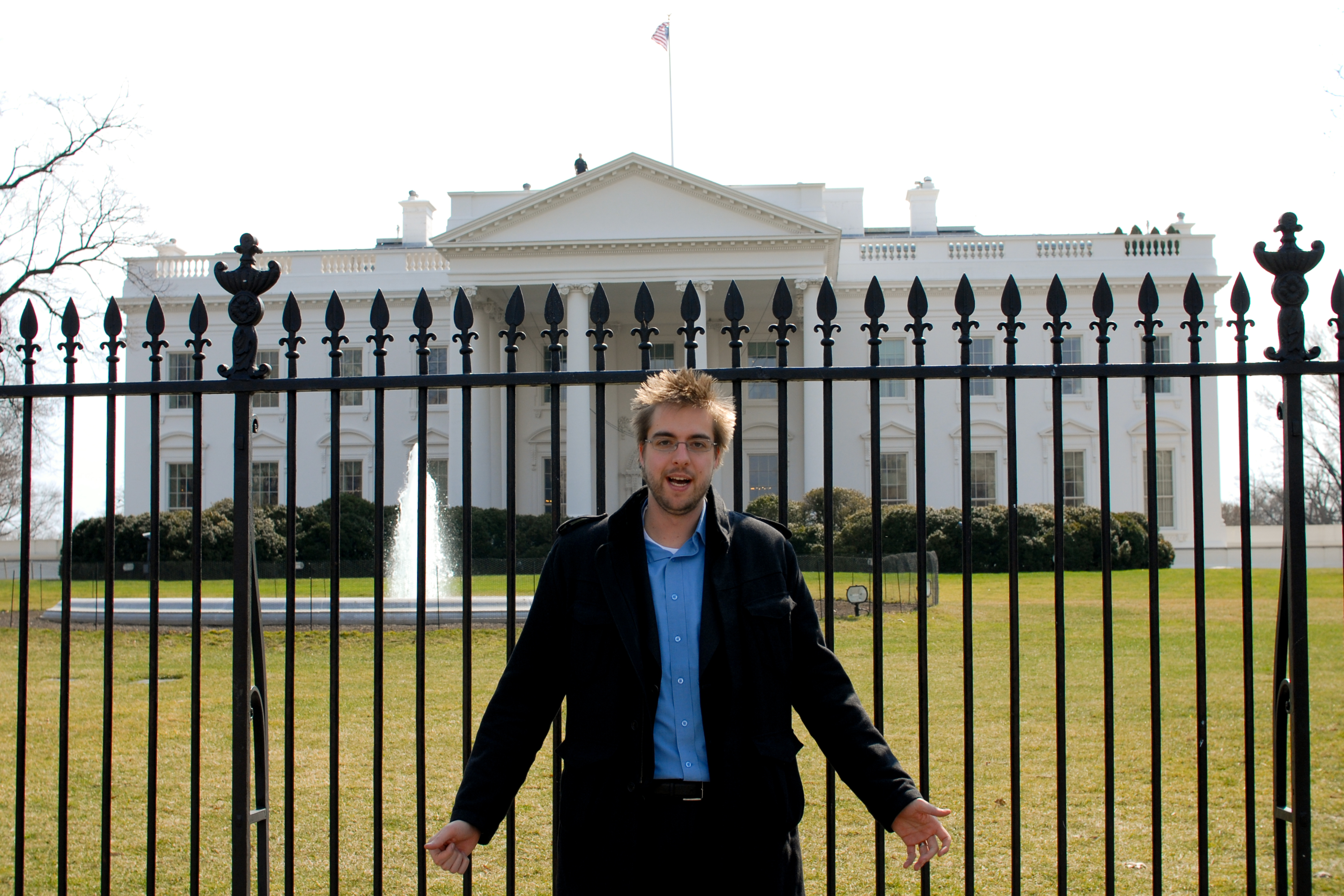 A man stands in front of a black fence with the White House visible behind him.