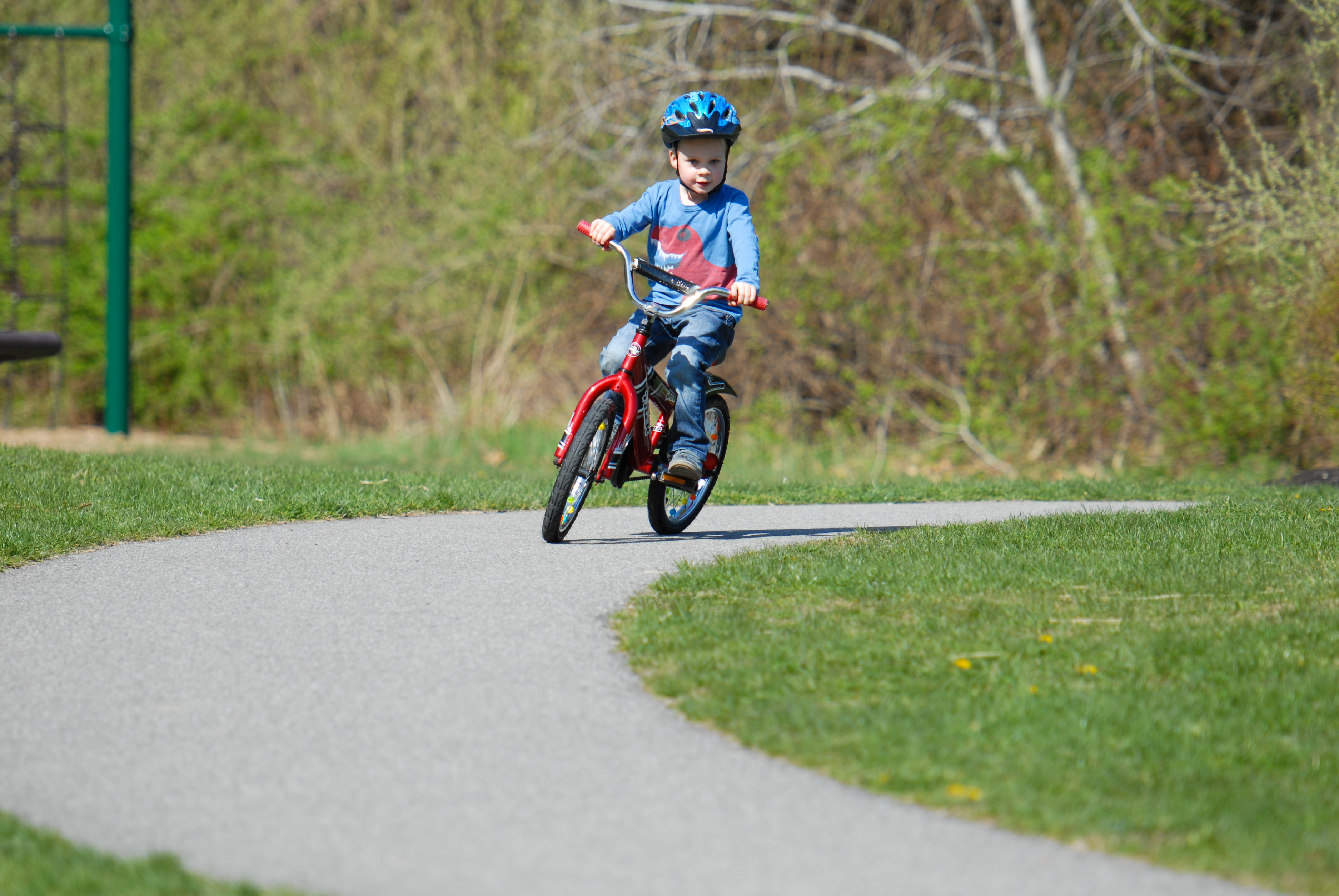 A young child rides a red bicycle without training wheels for the first time on a paved path.