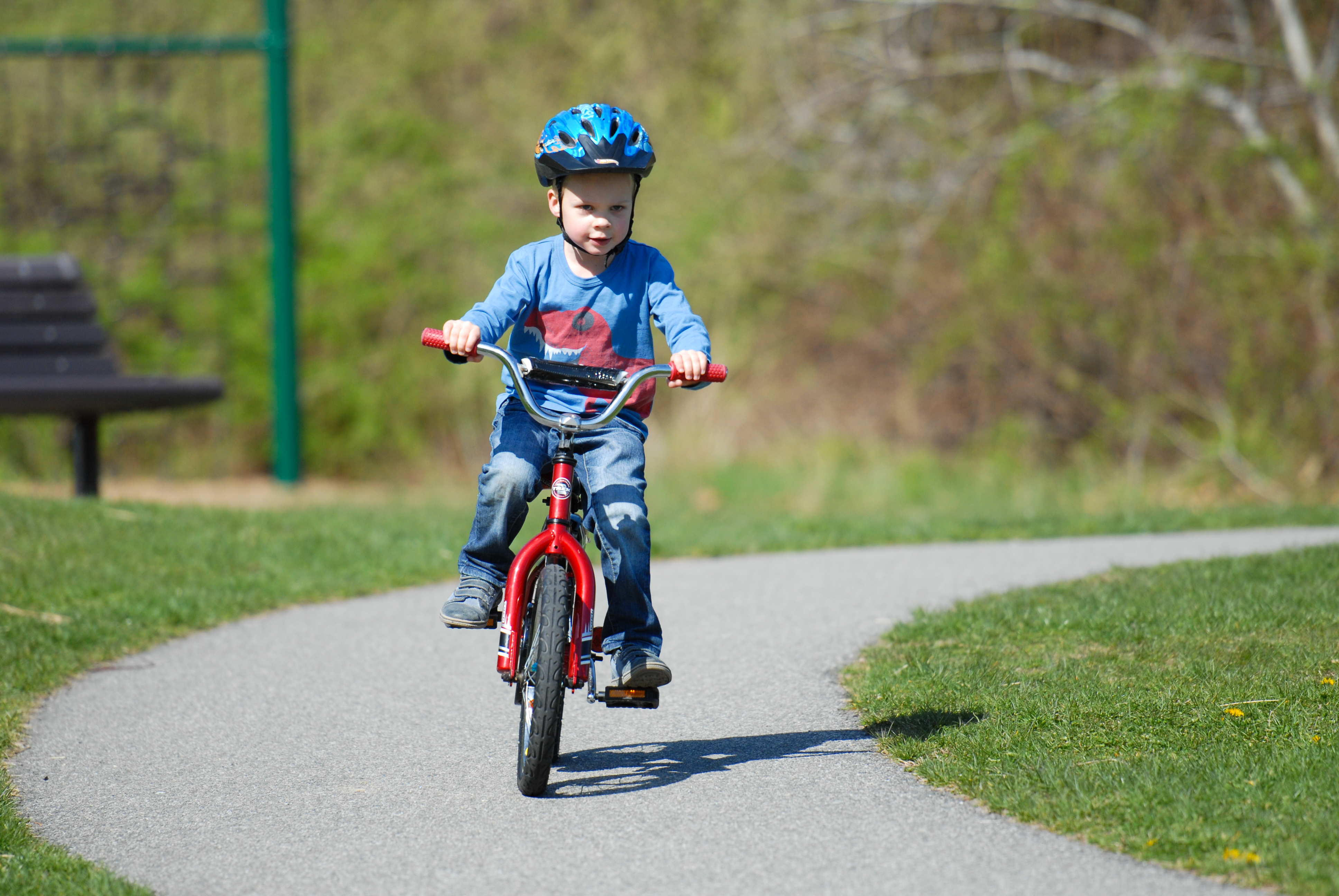 A young child rides a red bicycle without training wheels for the first time on a paved path.