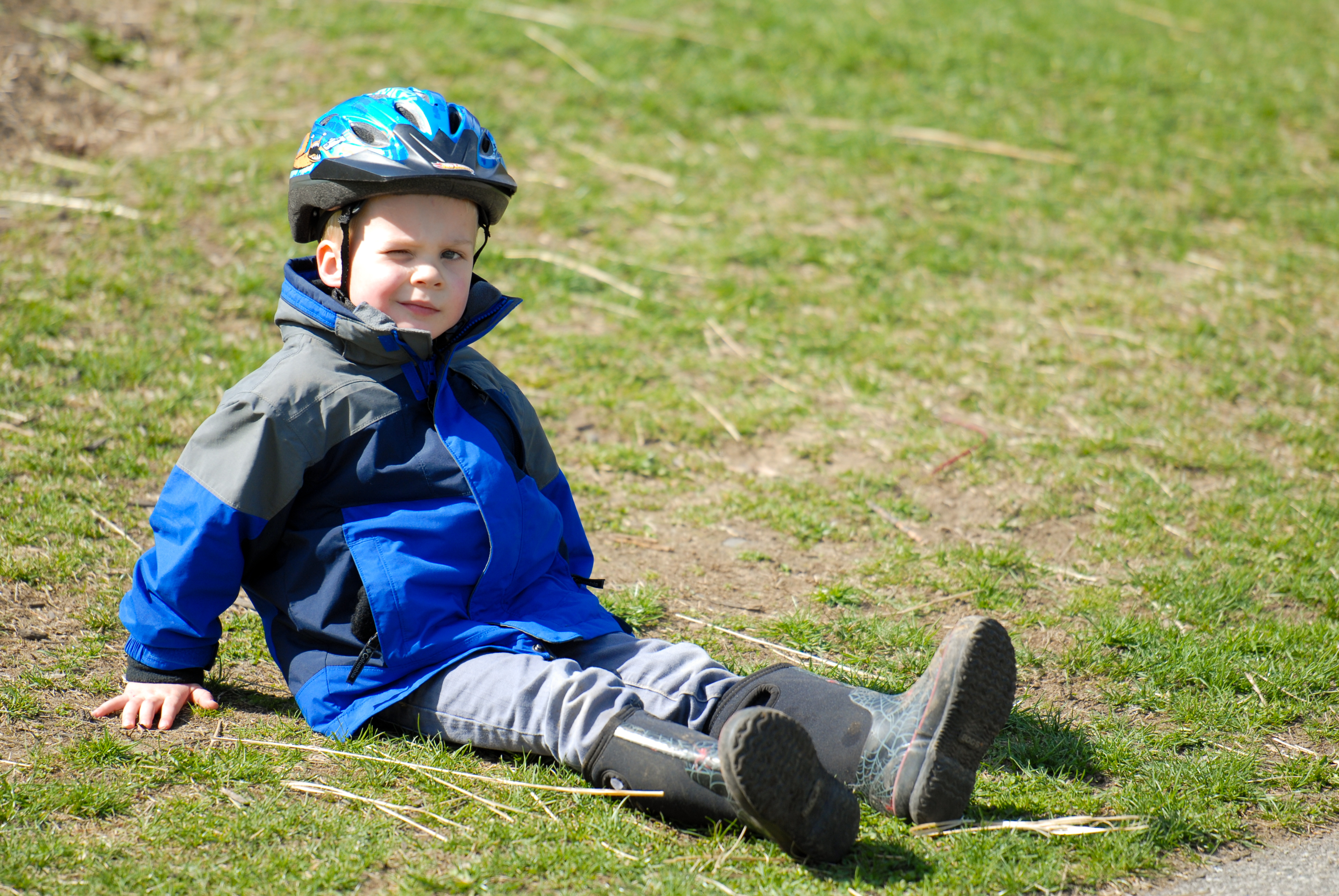 A child wearing a helmet and jacket sits on the grass, leaning back with legs stretched out.