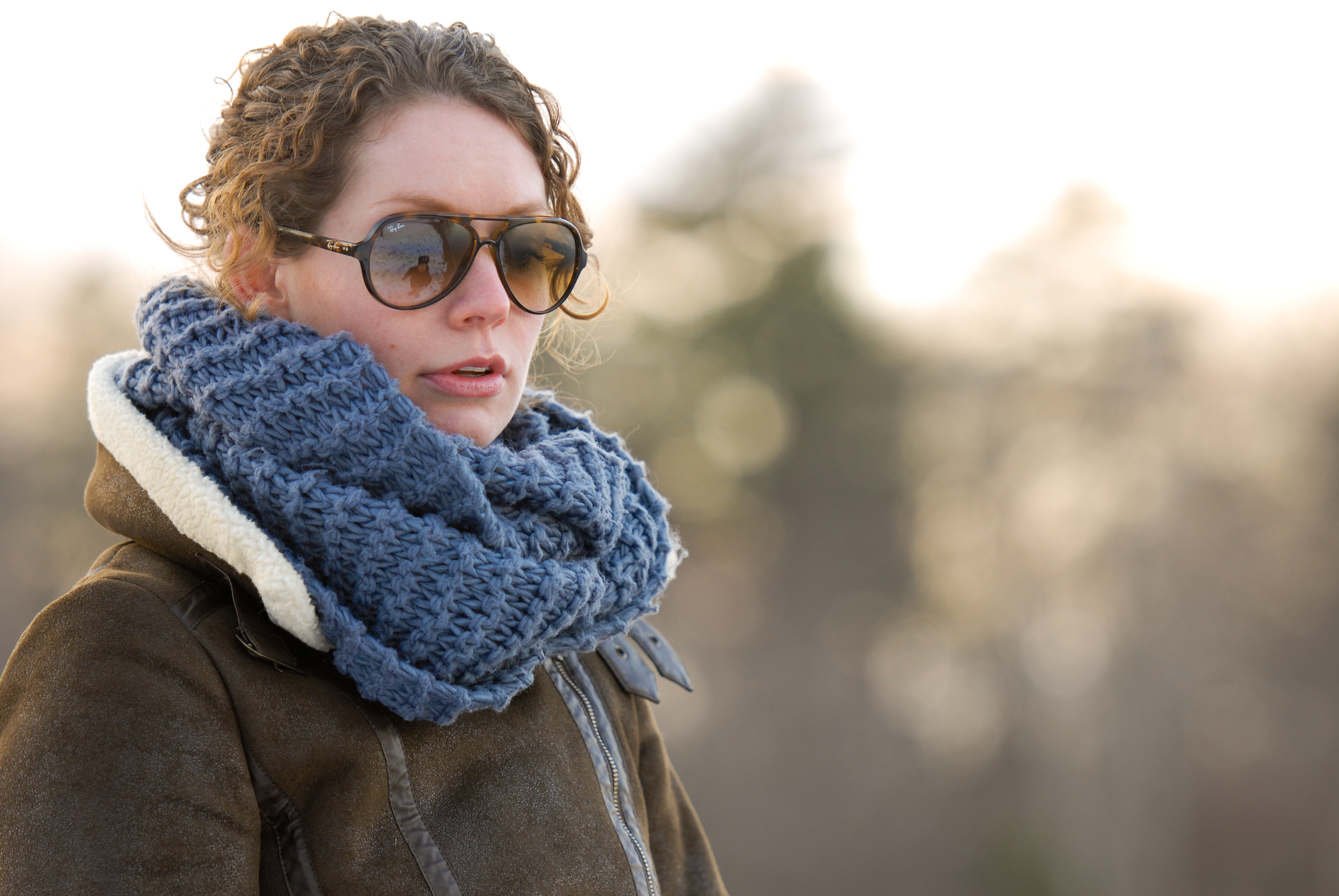 A person wearing sunglasses, a thick blue scarf, and a brown coat stands outdoors in cool weather.