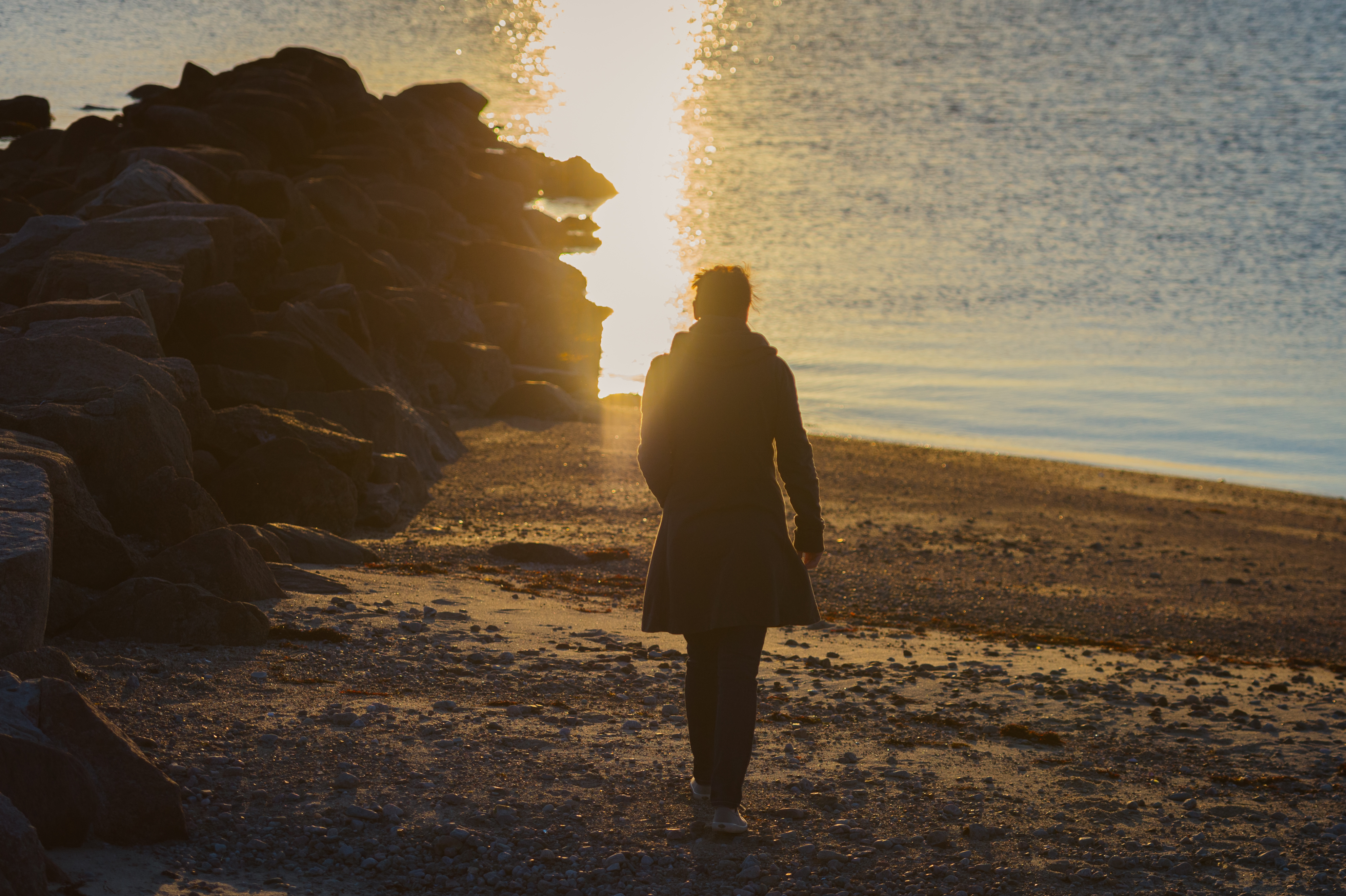 A person in a coat walks along a rocky shoreline toward the water, with sunlight reflecting on the surface.