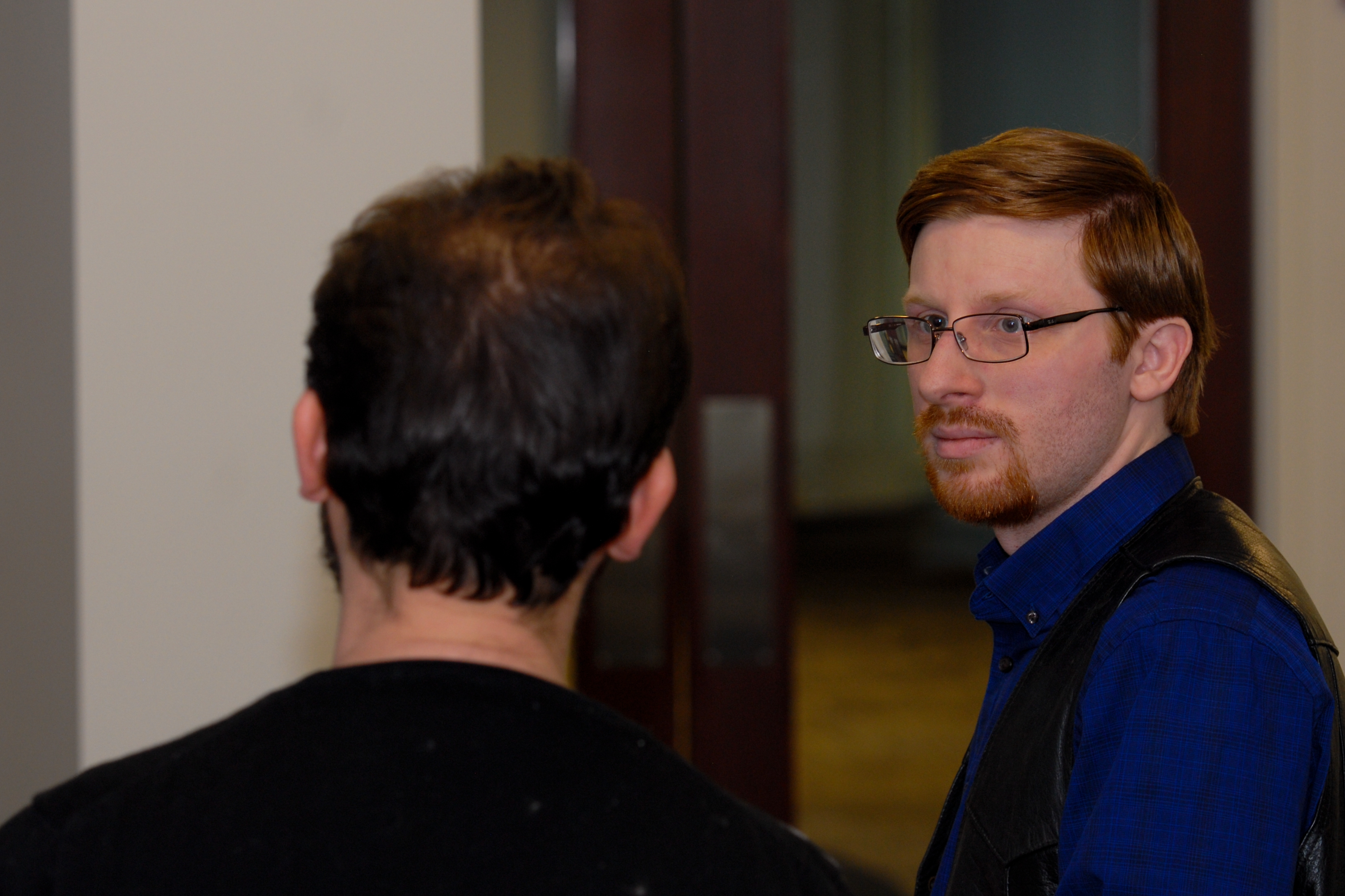 Two people in conversation indoors, one facing away from the camera, the other looking towards them with a serious expression.