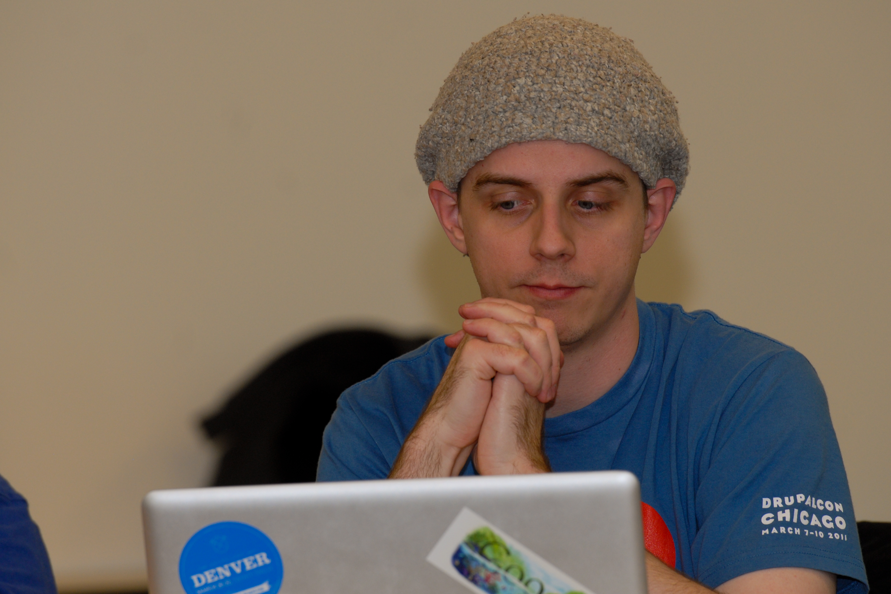 A person in a blue shirt and knit hat looks at a laptop with hands clasped together.